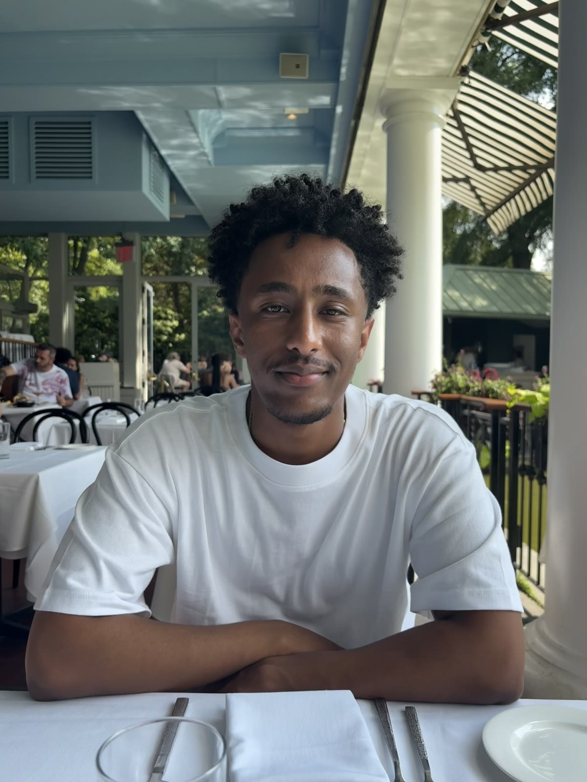 Binyam seated at an outdoor restaurant, wearing a white T-shirt.